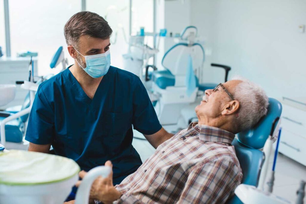 A male dentist wearing a face mask speaking with an older male patient in a dental office setting.