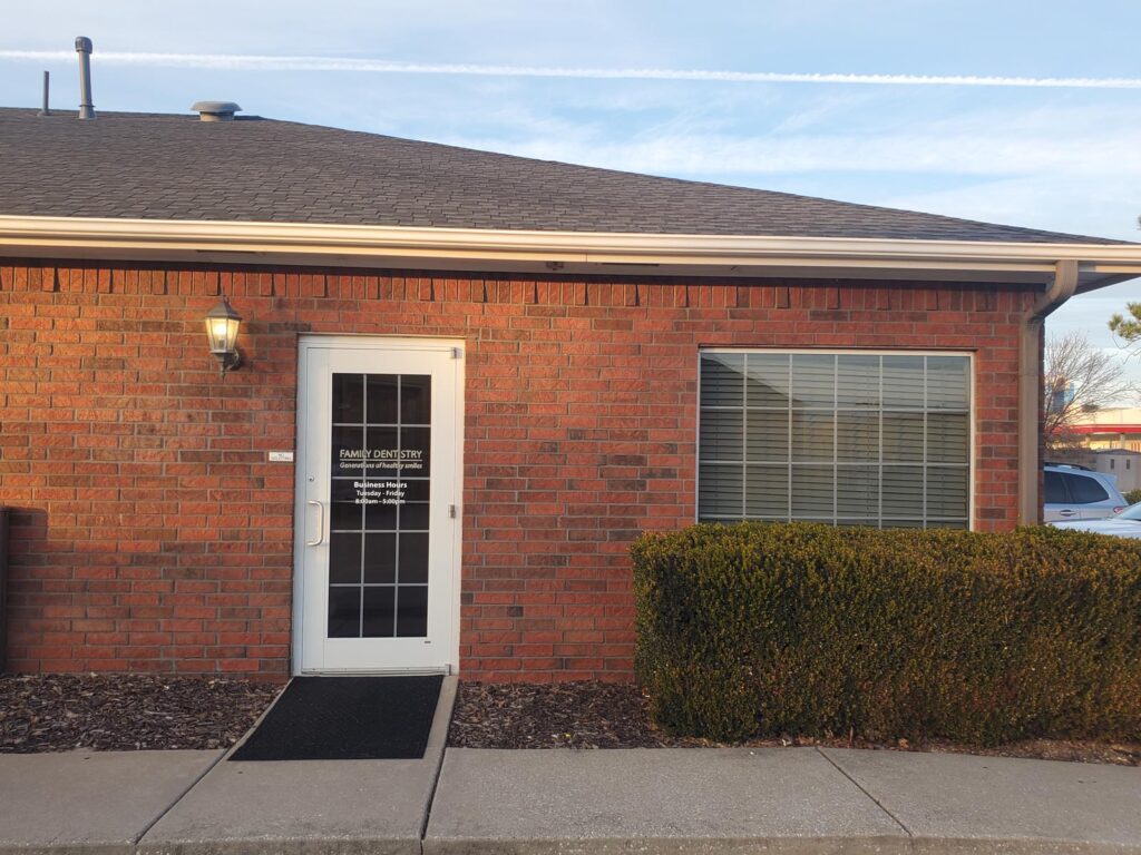 A single-story brick dental office with a glass door entrance and a large window, surrounded by a sidewalk and bushes.