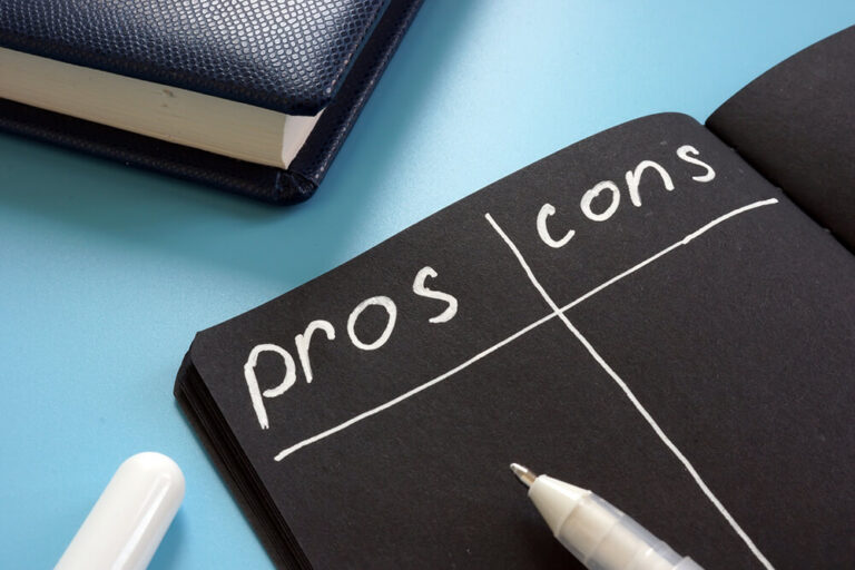 A black chalkboard with "pros" and "cons" written in white chalk, placed on a blue surface with a pen and notebook.